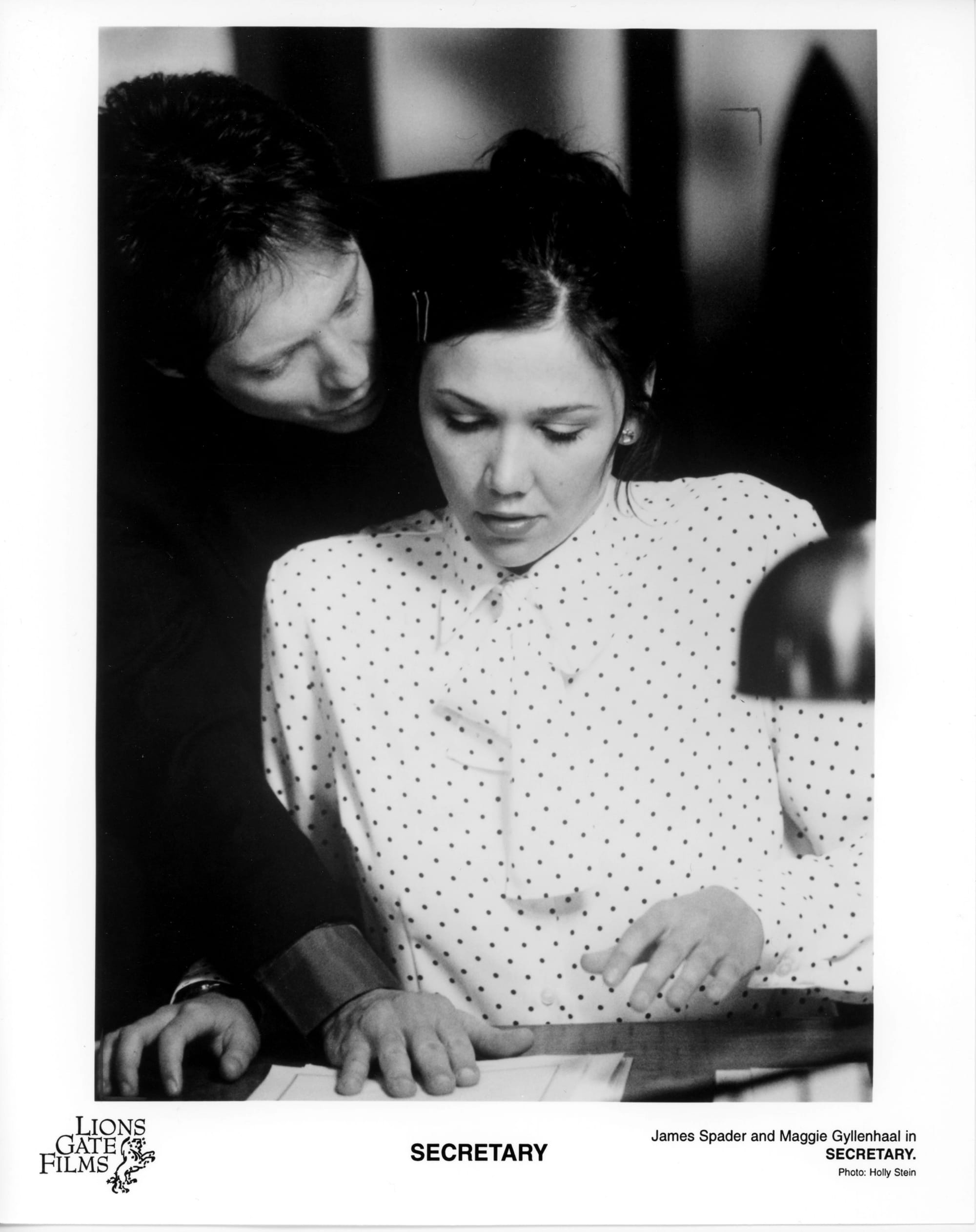 B&W Publicity Photo - A woman in a white blouse with tiny polka dots sits as a desk while a man in black leans over her with mouth close to ear.