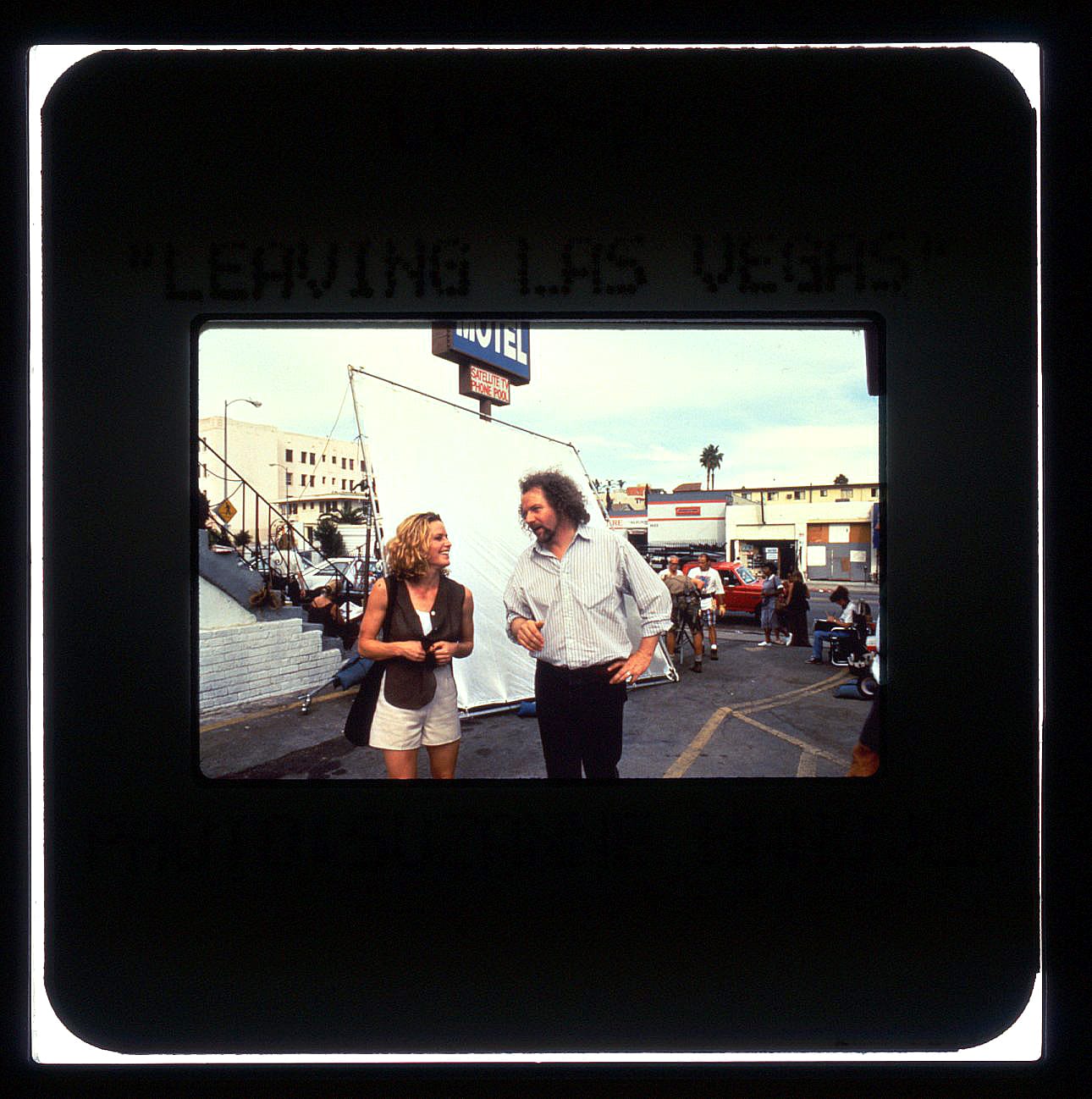 Color publicity slide of a woman standing and laughing while talking to a man in a motel parking lot. Behind them is a giant white sheet affixed tautly to a steel rod frame. Behind that is the motel sign.