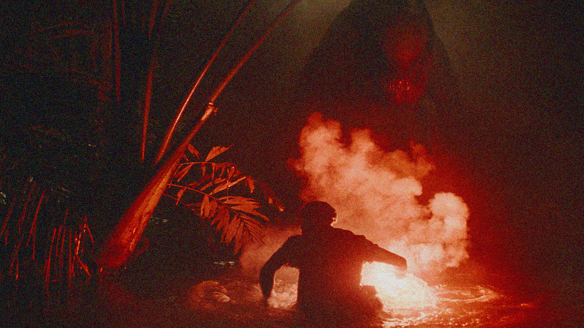 Red tinted smoky scene in the water with a man holding flare in the foreground and a giant monster dinosaur approaching in the background.