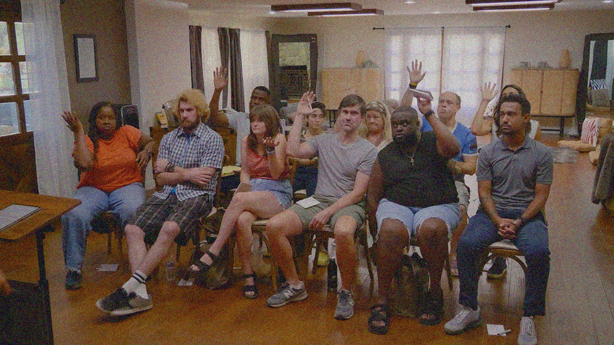 A group of people sit in a classroom-like setting on two rows of chairs. Some have their hands raised, others do not.
