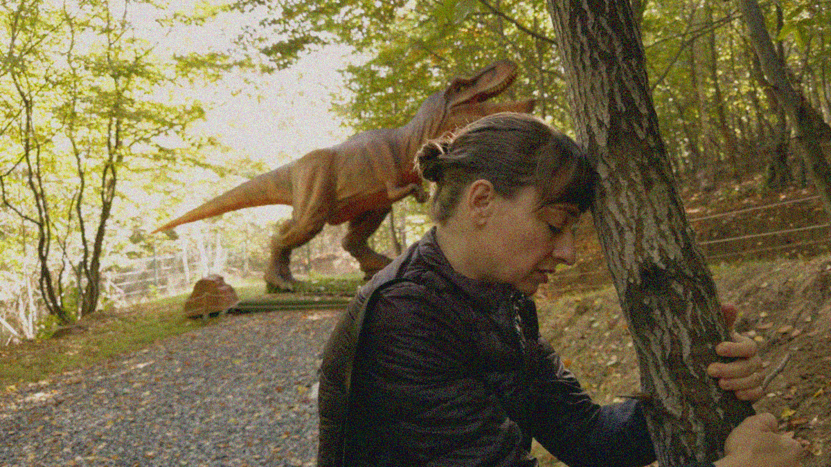 A woman with eyes closed says a prayer with forehead on tree trunk and arms around it. An animatronic dinosaur stands behind her in the park.