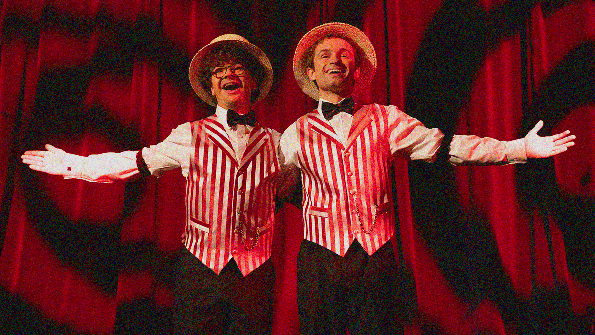 Two young men dressed like they're in a barbershop quartet with striped vests, bowties, and hats lock arms spread hands with a smile against a red stage curtain.