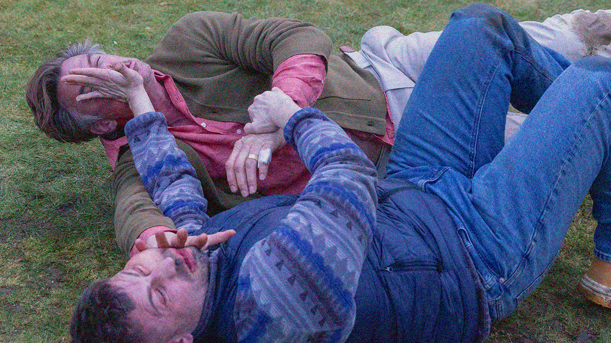 Two grown men lying on the grass fighting, each one as the other's hand in their face pushing it away.