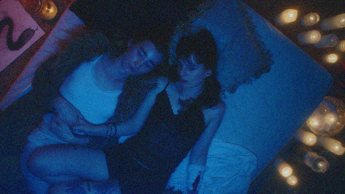 Overhead shot of two young women lying on their backs in bed that's tinted blue besides the yellow candles filling the right side of the frame.