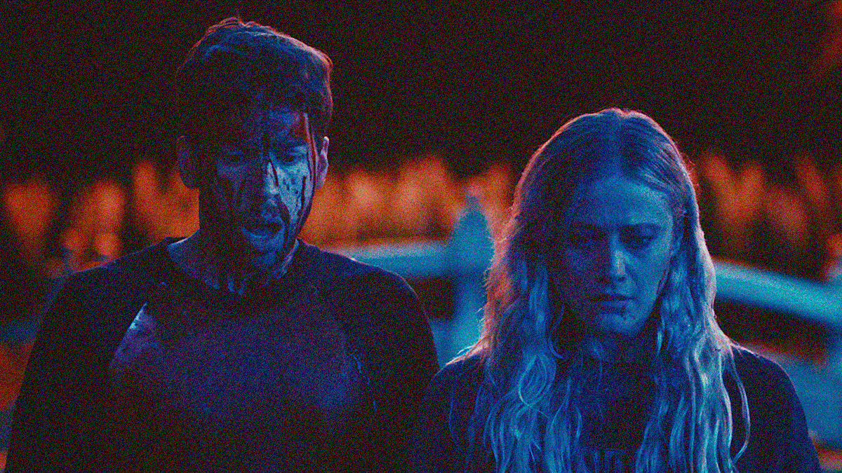 A man and woman look downwards while facing the camera. Both are shown from the chest up and covered in blood.