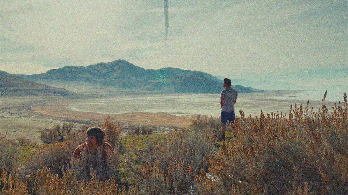 A long shot in nature with mountains in the distance and a long unnatural column coming down from the sky. In the foreground are two people relieving their bladders in the grass. She is squatting at left, he is standing at right.