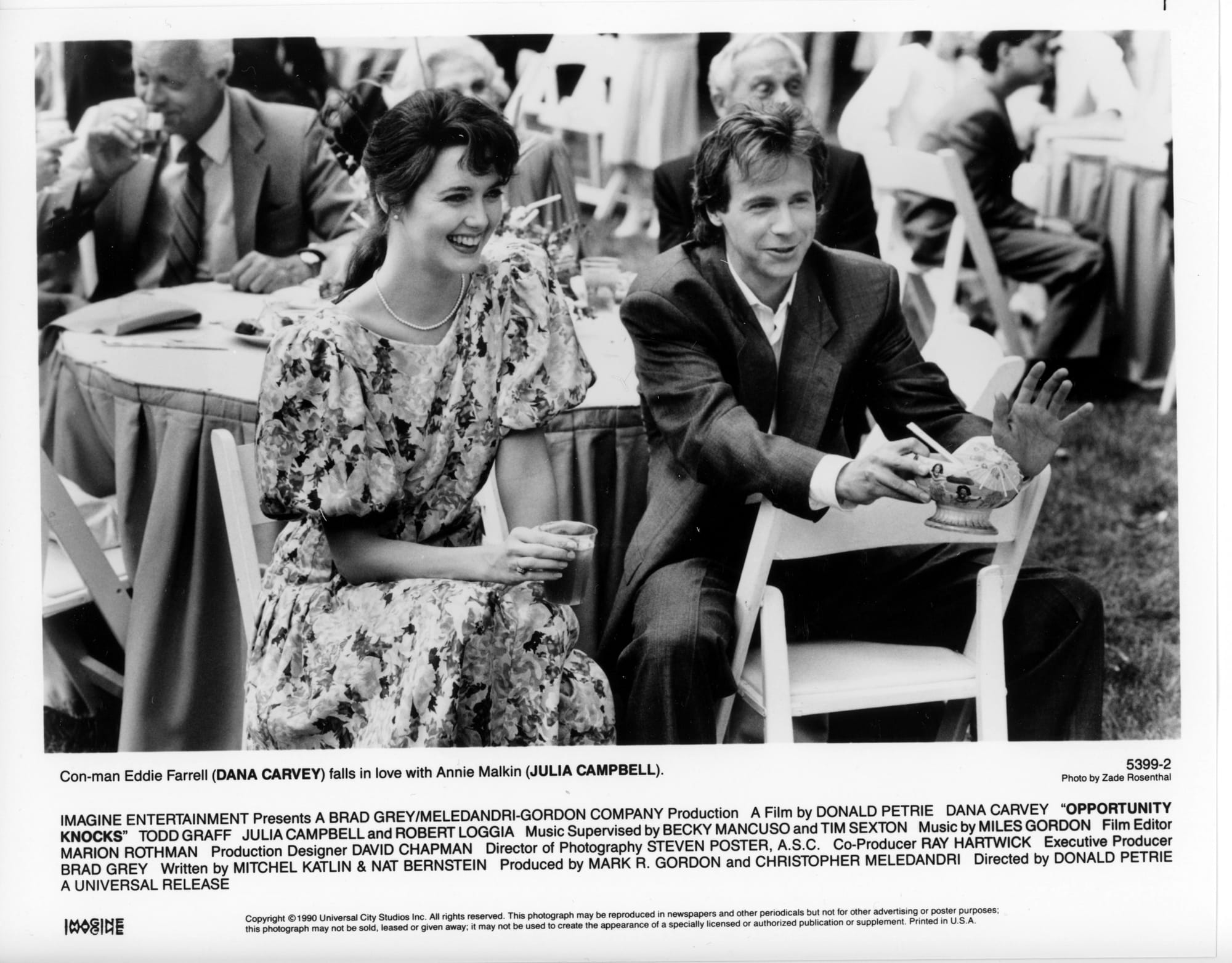 B&W Publicity Photo: A woman and man sit at a wedding reception table. She is facing forward and laughing with a drink in hand. He's facing forward, but sitting on his chair is backwards. He's holding a bowl with a paper umbrella.