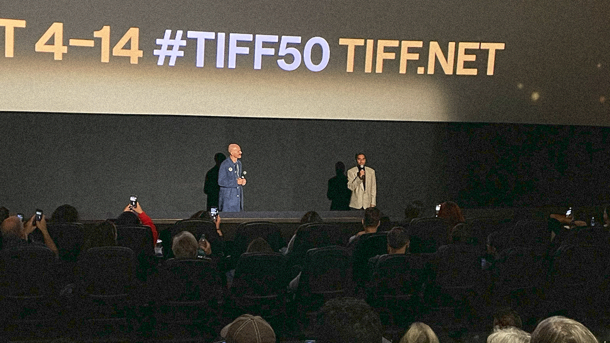 Two men with microphones standing in front of a full theater with the TIFF 50 brand info screen projected behind them.