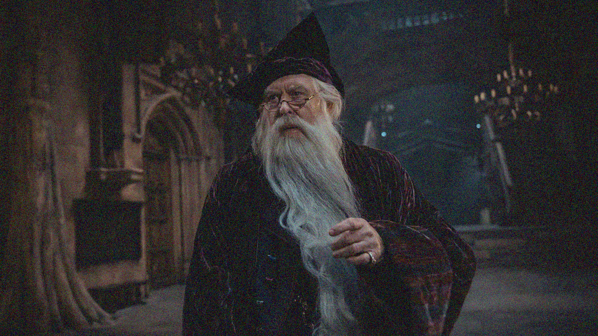 A long, white-bearded man in robe and hat with pinky ring stands in the foreground of a medieval-like mansion hall.