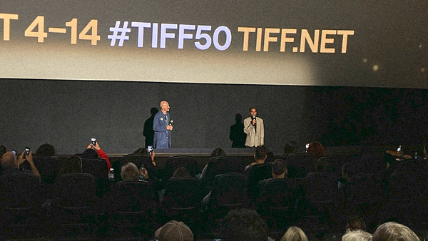 Two men with microphones standing in front of a full theater with the TIFF 50 brand info screen projected behind them.