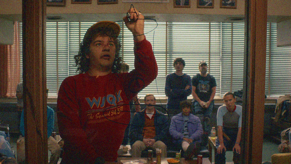 A teen boy in a red radio station sweatshirt draws on a window with black marker while a bunch of people sit behind him watching. We see it all through the glass.