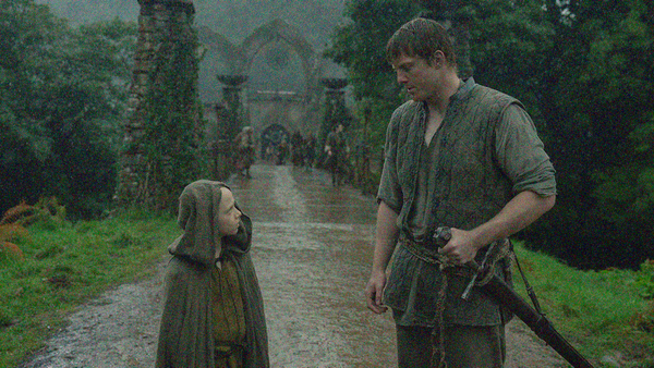 A small bald boy in a hooded cloak at left stands and looks at a large man at right looking down at him in the rain with left hand on sword hilt.